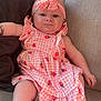 baby, child, couch, pink_dress, headband, strawberry_pattern, indoor, cute, infant, sitting, plush, comfortable, pout, young_child, human, portrait, relaxed, soft_fabric, home, adorable