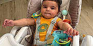Léo participe au concours pour gagner de l'argent avec cette photo : baby, high_chair, toy, kitchen, child, infant, orange_clothing, plastic_toy, flooring, cabinet, face, person, seatbelt, indoor, smiling, furniture, cute, sitting, young_child, play