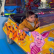 Léo participe au concours pour gagner de l'argent avec cette photo : child, toddler, amusement_ride, kiddie_car, carousel, colorful, outdoor, daylight, playground, cartoon_characters, yellow_car, blue_floor, face, person, clothing, fun, park, vehicle, smile, family