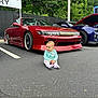 baby, child, car, red_car, parking_lot, asphalt, outdoor, trees, greenery, smiling, sitting, casual_clothing, sports_car, daylight, cute, person, vehicle, road, hood, license_plate