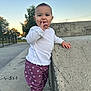 toddler, child, outdoor, concrete, pavement, clothing, white_shirt, purple_pants, flowers, shoes, hand, face, curious, sky, trees, daylight, standing, person, urban, casual