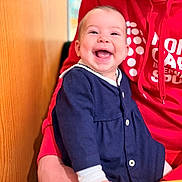Lou a rejoint le concours — aidez-le/la à gagner de superbes lots ! apparel, baby, child, closeup, clothing, cute, face, hands, happy, holding, indoor, infant, joyful, navy_blue, person, portrait, red_hoodie, sitting, smiling, young
