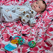 Alex is registered to the contest to win money with this photo: baby, infant, plush_toy, dinosaur_onesie, blanket, pink, white, floral_pattern, toy, rattle, lying_down, face, hand, cute, child, indoor, soft_toy, comfort, relaxed, young_child