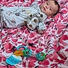 baby, infant, plush_toy, dinosaur_onesie, blanket, pink, white, floral_pattern, toy, rattle, lying_down, face, hand, cute, child, indoor, soft_toy, comfort, relaxed, young_child