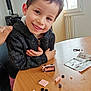 child, smile, table, toy, building_blocks, instruction_sheet, indoor, window, chair, clothing, person, happy, face, hands, wood, play, casual, hoodie, short_hair, daylight