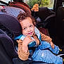 toddler, child, car_seat, curly_hair, blue_eyes, hand_gesture, casual_clothing, jeans, sneakers, car_interior, rainy_window, toy, smile, seatbelt, vest, sitting, indoor, happy, person, portrait