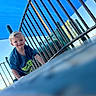 child, toddler, playground, climbing, blue_sky, metal_bars, outdoor, smiling, blonde_hair, tshirt, dinosaur_print, daylight, recreation, fun, play, young_child, happy, person, casual_clothing, park