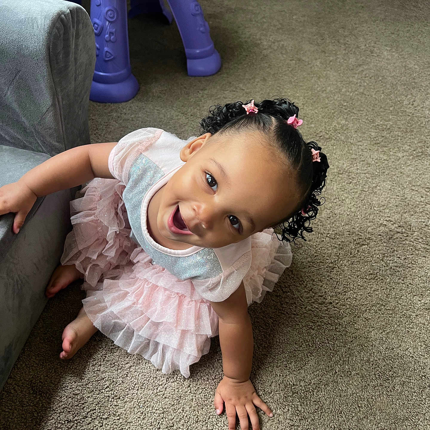 Kaylani is registered to the contest to win money with this photo: toddler, child, smiling, pink_dress, curly_hair, hair_clips, carpet, couch, toy_table, indoor, playful, happy, person, cute, portrait, baby, furniture, floor, home, bright