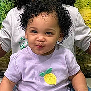 Kaylani is registered to the contest to win money with this photo: toddler, child, curly_hair, purple_shirt, lemon_patch, smiling, playful, person, portrait, indoor, adult_in_background, green_background, short_sleeves, cute, young_child, face, black_hair, happy, clothing, sitting