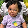 toddler, child, curly_hair, purple_shirt, lemon_patch, smiling, playful, person, portrait, indoor, adult_in_background, green_background, short_sleeves, cute, young_child, face, black_hair, happy, clothing, sitting