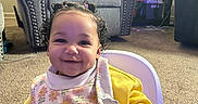 Kaylani joined the competition — help win amazing prizes! baby, child, high_chair, bib, yellow_clothing, curly_hair, pigtails, smile, indoor, living_room, carpet, armchair, lamp, furniture, happy, person, portrait, cute, infant, seated