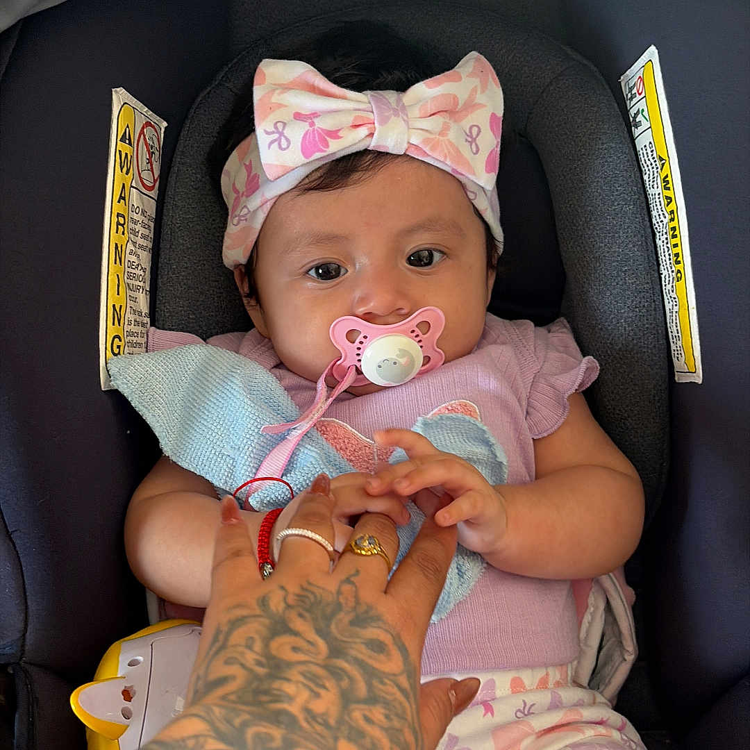 Yolanda is registered to the contest to win money with this photo: baby, child, infant, pacifier, headband, car_seat, tattoo, hand, pink_clothing, patterned_pants, blanket, indoor, cute, holding_hand, person, seated, closeup, soft_lighting, comfortable, adorable