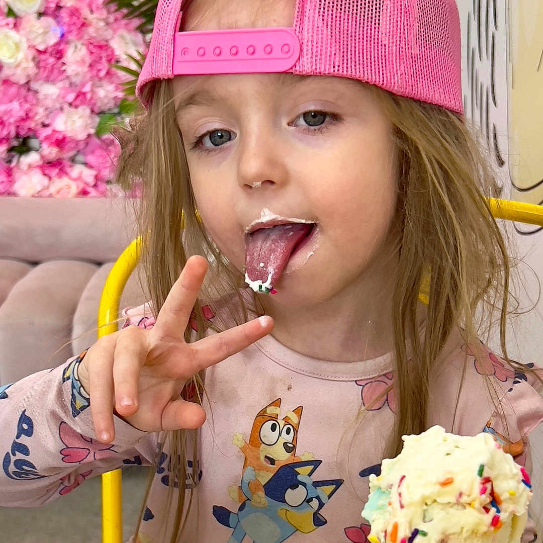 Jocyne is registered to the contest to win money with this photo: cartoon_print, child, colorful, cone, floral_background, fun, girl, happy, ice_cream, indoor, long_hair, messy_face, peace_sign, pink_cap, pink_dress, playful, sprinkles, sweet, tongue_out, yellow_chair