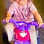 child, girl, smiling, toy_bike, indoor, blonde_hair, pink_shirt, cartoon_print, slippers, happy, riding, floor, play, cute, small_bike, person, young_child, fun, joy, casual_clothing