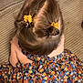 child, hair, hair_bun, hair_accessory, yellow_flower, dress, floral_pattern, colorful_clothing, sitting, top_view, brown_hair, childrens_fashion, indoors, textured_surface, patterned_fabric, cute, young_child, hairstyle, flower_clip, skin