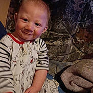 Elijah is registered to the contest to win money with this photo: baby, child, smiling, portrait, sitting, indoor, blanket, bed, pajamas, striped_clothing, patterned_clothing, face, hands, happy, cozy, home, furniture, fabric, warm_lighting, photograph