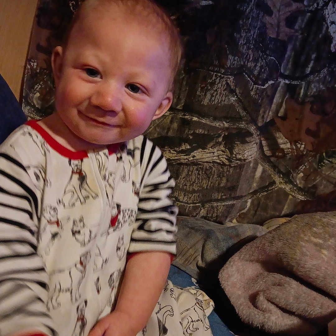 Elijah is registered to the contest to win money with this photo: baby, child, smiling, portrait, sitting, indoor, blanket, bed, pajamas, striped_clothing, patterned_clothing, face, hands, happy, cozy, home, furniture, fabric, warm_lighting, photograph