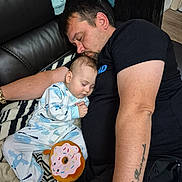 Luca Nicolae is registered to the contest to win money with this photo: baby, man, sleeping, couch, blanket, plush_toy, donut_toy, tattoo, watch, pajamas, black_tshirt, floor, wood_floor, child, indoor, rest, nap, family, bonding, casual_clothing