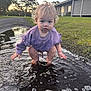 toddler, child, puddle, water, outdoor, grass, curly_hair, purple_sweater, squatting, playful, sunset, pathway, bubbles, building, nature, reflection, person, young_child, casual_clothing, daylight