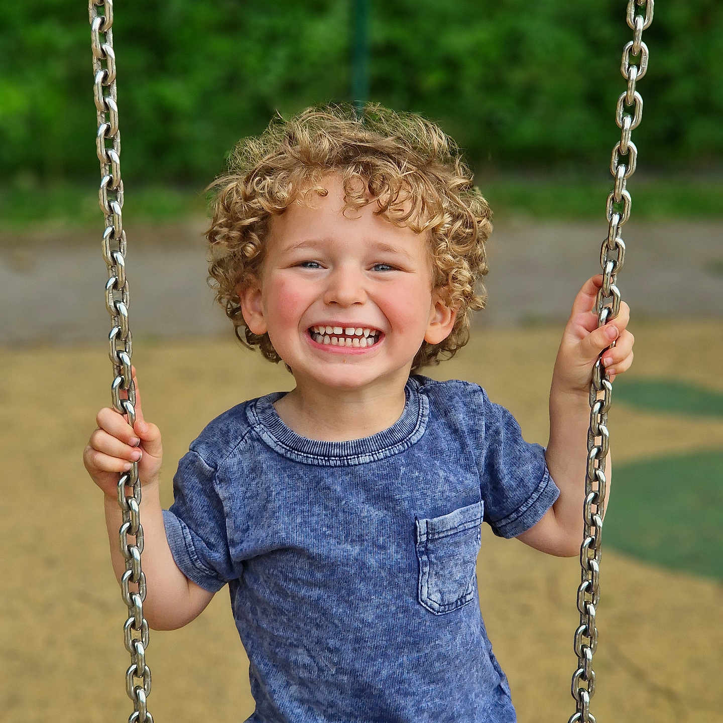 Jules participe au concours pour gagner de l'argent avec cette photo : blue_shirt, boy, casual_clothes, child, curly_hair, daylight, face, fun, happy, holding, joy, metal_chain, outdoor, park, playground, recreation, short_sleeves, smiling, swing, toddler
