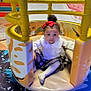 child, colorful, curly_hair, cute, floor_mat, fun, girl, indoor_play_area, looking_at_camera, padded, plaid_skirt, play_structure, playground, portrait, red_bow, sitting, soft_expression, toddler, white_blouse, young_child