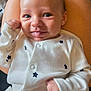 baby, infant, onesie, white_clothing, stars_pattern, tongue_out, smiling, face, hands, skin, person, cute, young_child, sitting, lap, happy, playful, indoor, closeup, portrait