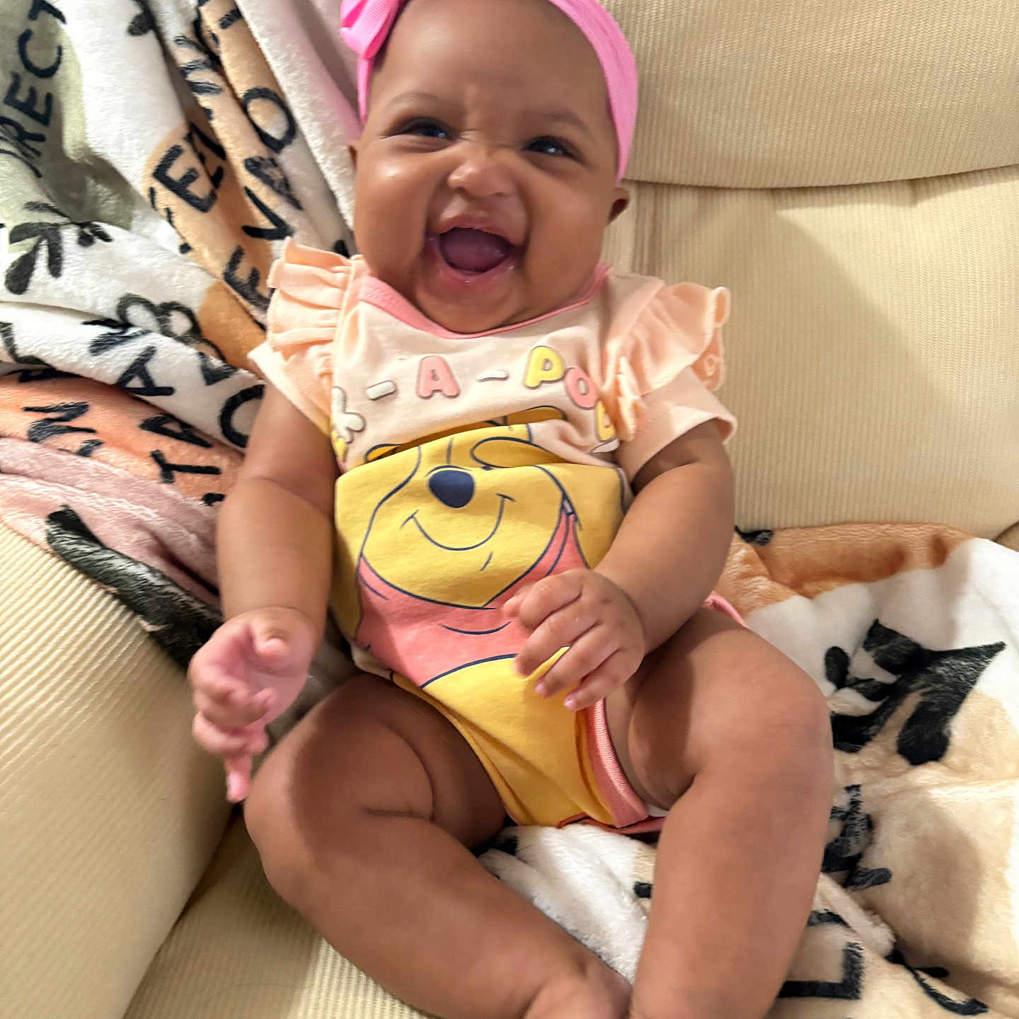 Kadence is registered to the contest to win money with this photo: baby, child, smiling, pink_headband, onesie, winnie_the_pooh, couch, blanket, happy, infant, sitting, cute, indoors, portrait, furniture, soft_toys, cozy, playful, small_baby, cute_expression