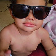 Royalty is registered to the contest to win money with this photo: baby, child, curly_hair, sunglasses, portrait, indoors, seated, close_up, face, lips, skin, infant, fashion, funny, looking_at_camera, blanket, red_fabric, purple_clothing, home, photography