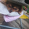 dog, small_dog, sleepy, hat, houndstooth, towel, cozy, resting, lying_down, brown_fur, face, eyes, nose, tongue, outdoor, patio, flowers, chair, textile, portrait