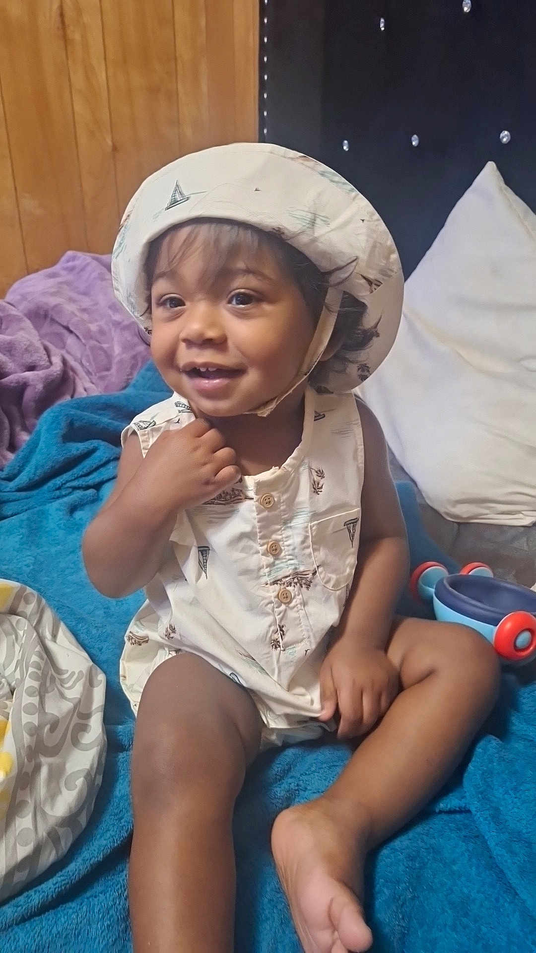Josiah is registered to the contest to win money with this photo: toddler, child, smiling, hat, sleeveless_shirt, indoor, blanket, pillow, toy, happy, cute, skin, foot, hand, face, sitting, wood_paneling, fabric, comfort, playful
