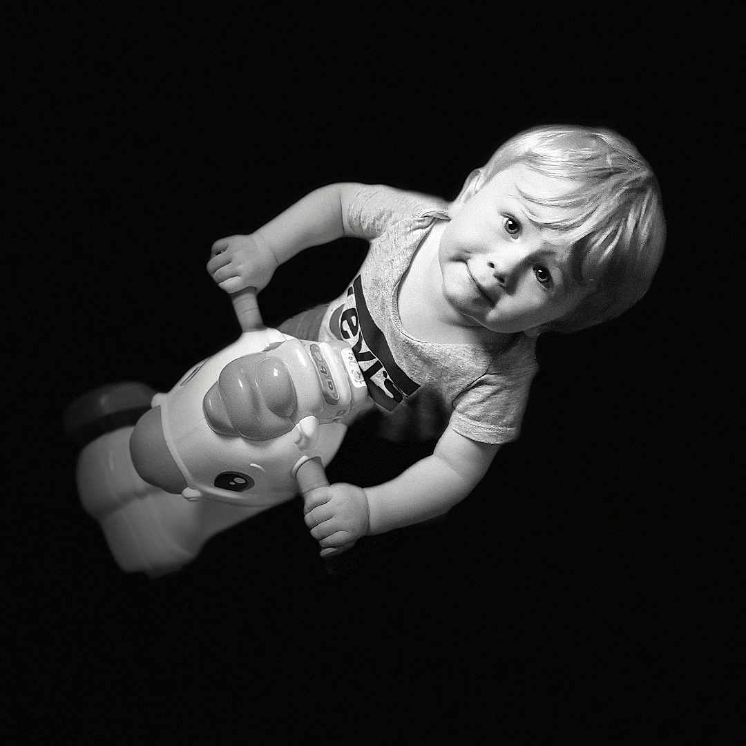 Dayvenn participe au concours pour gagner de l'argent avec cette photo : black_background, blond_hair, child, curious, cute, floating, fun, happy, indoor, isolated, monochrome, playful, portrait, riding, short_sleeves, smiling, toddler, toy, unicorn, young_child