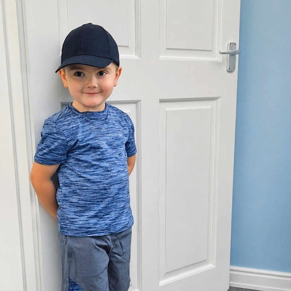 Corey joined the competition — help win amazing prizes! blue_shirt, boy, cap, casual_clothing, child, feet, floor, happy, indoor, light, person, portrait, room, shorts, smile, sneakers, socks, standing, wall, white_door