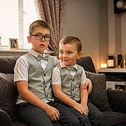 Corey is registered to the contest to win money with this photo: children, brothers, portrait, sitting, sofa, living_room, formalwear, vest, bow_tie, glasses, smiling, indoors, family, siblings, young, boy, furniture, home, warm_lighting, photography