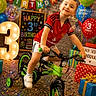 child, birthday, balloons, bike, confetti, presents, celebration, smiling, happy, red_shirt, indoor, party, decorations, number_3, training_wheels, colorful, young_boy, festive, floor, lighting