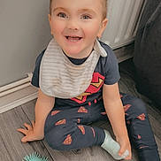 Corey joined the competition — help win amazing prizes! toddler, child, smiling, pajamas, superman_logo, bib, toy, floor, wooden_floor, indoor, happy, sitting, cute, playful, person, young_child, short_hair, sock, home, radiator