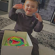 Corey is registered to the contest to win money with this photo: birthday, box, cake, carpet, celebration, child, cute, dinosaur, floor, happy, home, indoor, living_room, person, shoes, smile, sweatshirt, toddler, toy, young