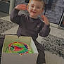 Corey is registered to the contest to win money with this photo: birthday, box, cake, carpet, celebration, child, cute, dinosaur, floor, happy, home, indoor, living_room, person, shoes, smile, sweatshirt, toddler, toy, young