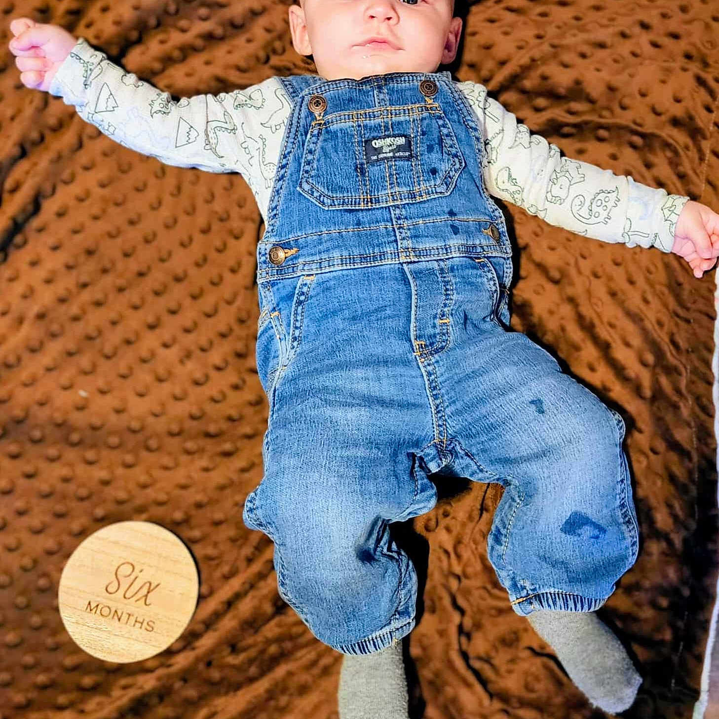 Kaspian joined the competition — help win amazing prizes! arms_outstretched, baby, brown_blanket, casual, child, clothing, cute, denim_overalls, dinosaur_print, face, indoor, infant, long_sleeve_shirt, lying_down, portrait, six_months, socks, soft_blanket, texture, wooden_sign