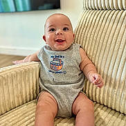 Amir is registered to the contest to win money with this photo: baby, child, infant, smiling, happy, portrait, sitting, armchair, furniture, home, indoor, onesie, clothing, bare_legs, socks, face, eyes, short_hair, cozy, text_on_clothing