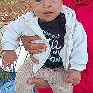 Layan a rejoint le concours — aidez-le/la à gagner de superbes lots ! baby, child, person, adult_hand, white_jacket, beige_pants, colorful_shoes, red_hoodie, blue_chair, outdoor, fence, table, curious_expression, sitting, holding, portrait, casual_clothing, daylight, close_up, cute