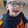 child, glasses, tongue_out, superman_logo, hoodie, indoor, smiling, playful, blond_hair, face, person, sitting, table, drawing, paper, colorful, chair, young_child, casual_clothing, happy