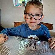Timéo a rejoint le concours — aidez-le/la à gagner de superbes lots ! child, boy, glasses, table, indoor, window, greenery, smile, blue_shirt, hands, face, portrait, seated, natural_light, background, wooden_chair, curtain, casual, home, young