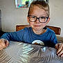child, boy, glasses, table, indoor, window, greenery, smile, blue_shirt, hands, face, portrait, seated, natural_light, background, wooden_chair, curtain, casual, home, young