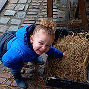 Maïdine a rejoint le concours — aidez-le/la à gagner de superbes lots ! child, curly_hair, blue_jacket, rain_boots, straw, black_container, cobblestone_floor, wet_floor, wooden_table, smiling, outdoor, playful, person, curly_bun, muddy, rustic, crouching, happy, casual_clothing, daylight
