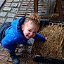 child, curly_hair, blue_jacket, rain_boots, straw, black_container, cobblestone_floor, wet_floor, wooden_table, smiling, outdoor, playful, person, curly_bun, muddy, rustic, crouching, happy, casual_clothing, daylight