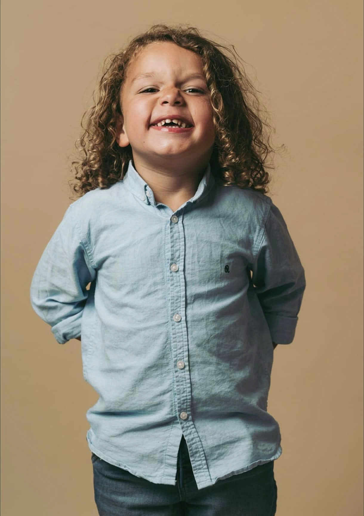 Maïdine a rejoint le concours — aidez-le/la à gagner de superbes lots ! child, curly_hair, smiling, gap_tooth, blue_shirt, casual_clothing, standing, portrait, happy, cute, kid, button_up, long_sleeves, jeans, hands_behind_back, studio, beige_background, light, person, fashion