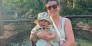 Daniel is registered to the contest to win money with this photo: baby, woman, child, smile, outdoor, fence, hat, sunglasses, topiary, greenery, sunlight, nature, shorts, tank_top, accessory, water, daytime, happy, person, portrait
