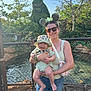baby, woman, child, smile, outdoor, fence, hat, sunglasses, topiary, greenery, sunlight, nature, shorts, tank_top, accessory, water, daytime, happy, person, portrait