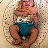baby, infant, smiling, bib, baby_seat, clothing, happy, cute, child, person, indoor, footwear, legs, face, skin, sitting, apparel, text_on_clothing, orange, blue_shorts