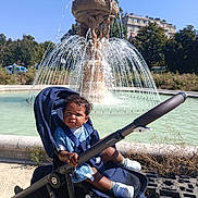 Noah a rejoint le concours — aidez-le/la à gagner de superbes lots ! child, toddler, stroller, fountain, water, outdoor, sunny, daylight, urban, park, trees, grass, baby_clothing, shoes, curly_hair, expression, person, seat, wheel, bottle